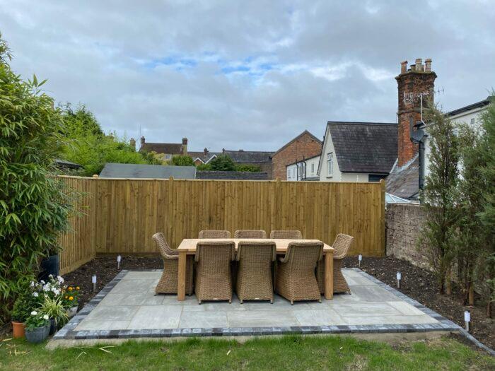 A backyard patio with a wooden table and six wicker chairs sits on Kandla Grey Sandstone Paving Slabs from Brisks. The space is enclosed by a wooden fence, and there is greenery and plants around the edge. Houses with brick chimneys are visible in the background under a cloudy sky, accented with subtle grey hues.