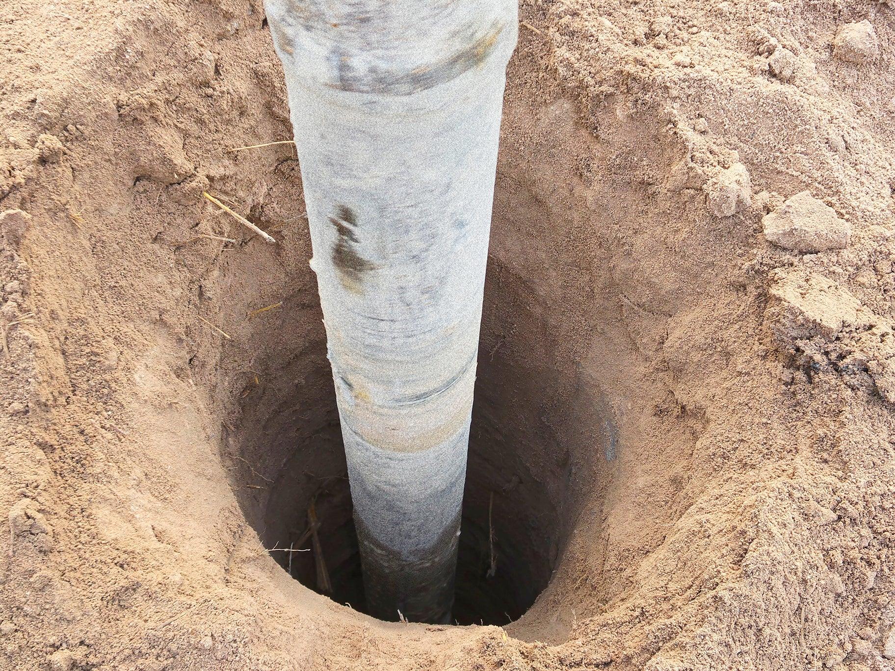 A close-up image of a metal pole partially buried in a hole dug in dry soil. The surrounding ground has an uneven, rough texture with some scattered small rocks and debris. The pole is upright with visible dirt and scratches on its surface, showcasing the effect of compressive strength on durable structures built with Brisks High Performance Concrete.