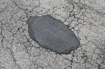 A close-up of a pothole that has been patched with a fresh layer of asphalt using Brisks Cold-Lay Patching Tarmac. The surrounding road surface shows significant cracking and wear, indicating that the area has experienced considerable damage over time, necessitating temporary road repair.