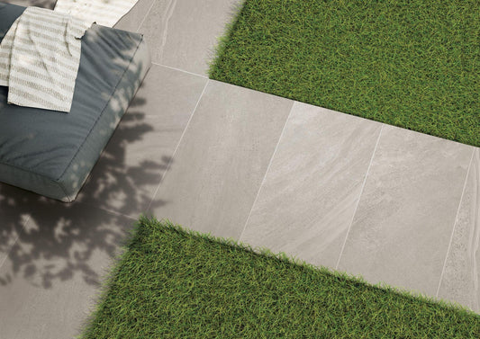 A modern outdoor setting featuring large rectangular Britstone Grey Porcelain Paving Slabs by Brisks forming a path amidst neatly trimmed grass. A cushioned seating area with a textured blanket rests on the tiles, and sunlight casts shadows of nearby foliage onto the slip-resistant surface.