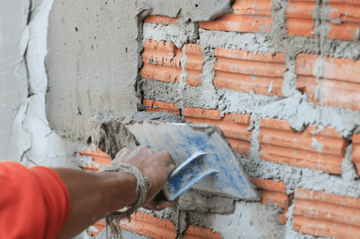 Close-up of a person's hand spreading a mortar mix on a brick wall using a trowel. The orange bricks are gradually being coated with Brisks Plastering Sand, smoothing the surface as they work.