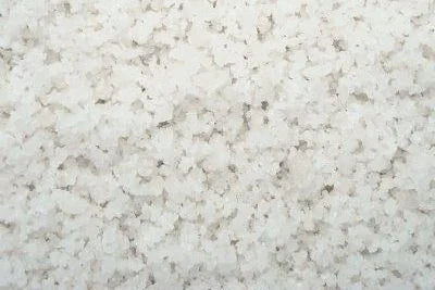 A close-up of coarse sea salt granules, displaying their crystalline texture and slightly varied sizes. The granules are white with a few small grayish specks scattered throughout. Reminiscent of Brisks' premium White De-Icing Salt, the overall appearance is rough and grainy.