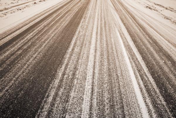 A road covered in light snow with visible tire tracks running parallel along its length. The surrounding ground also shows signs of snow, and the overall scene suggests a cold, wintery environment treated with Brisks White De-Icing Salt for a residue-free finish.